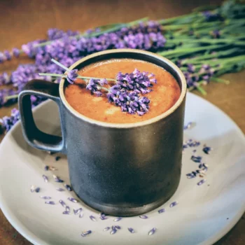 Brown mug of hot chocolate with lavender sprigs, sitting on grey plate with lavender bud and lavender bouquet in background