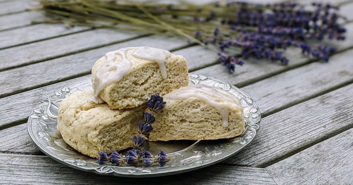 three Lavender Lemon Scones with lavender glaze, arranged on a silver plate that sits on a rustic wood table with dried lavender in background
