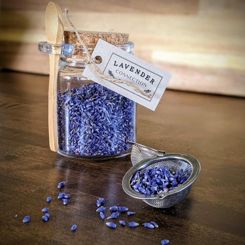 glass jar of culinary lavender bud, culinary lavender bud in open tea ball strainer spilling out, on wood counter with wood cutting board in background
