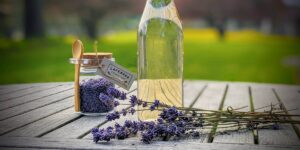 glass jar of culinary lavender, glass bottle of lavender simple syrup, lavender sprigs in front, all sitting on a wood table with farm grounds out of focus in background