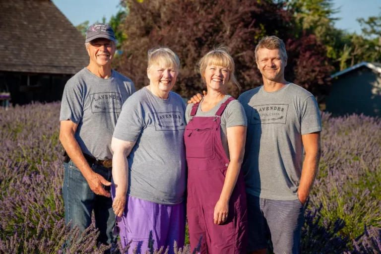 4 members of the Olson Family of Lavender Connection family farm close up, posing in lavender field with barn and trees in background